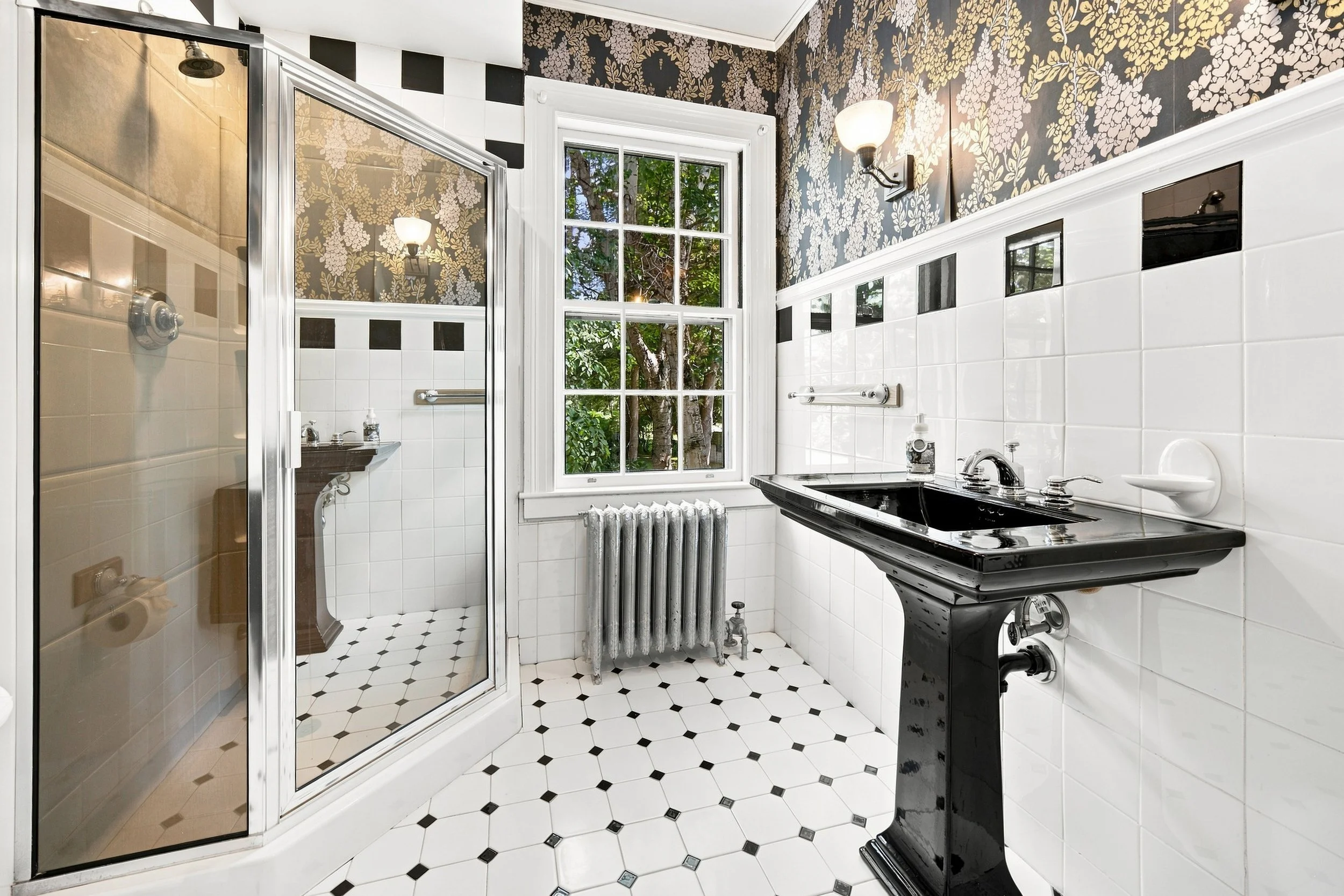 A bathroom with black and white diamond-patterned tile floor, white tiled walls with black accents, a window showing an outdoor view of trees, a black vintage sink, a radiator, a shower with a glass door, and floral wallpaper in the upper part of the