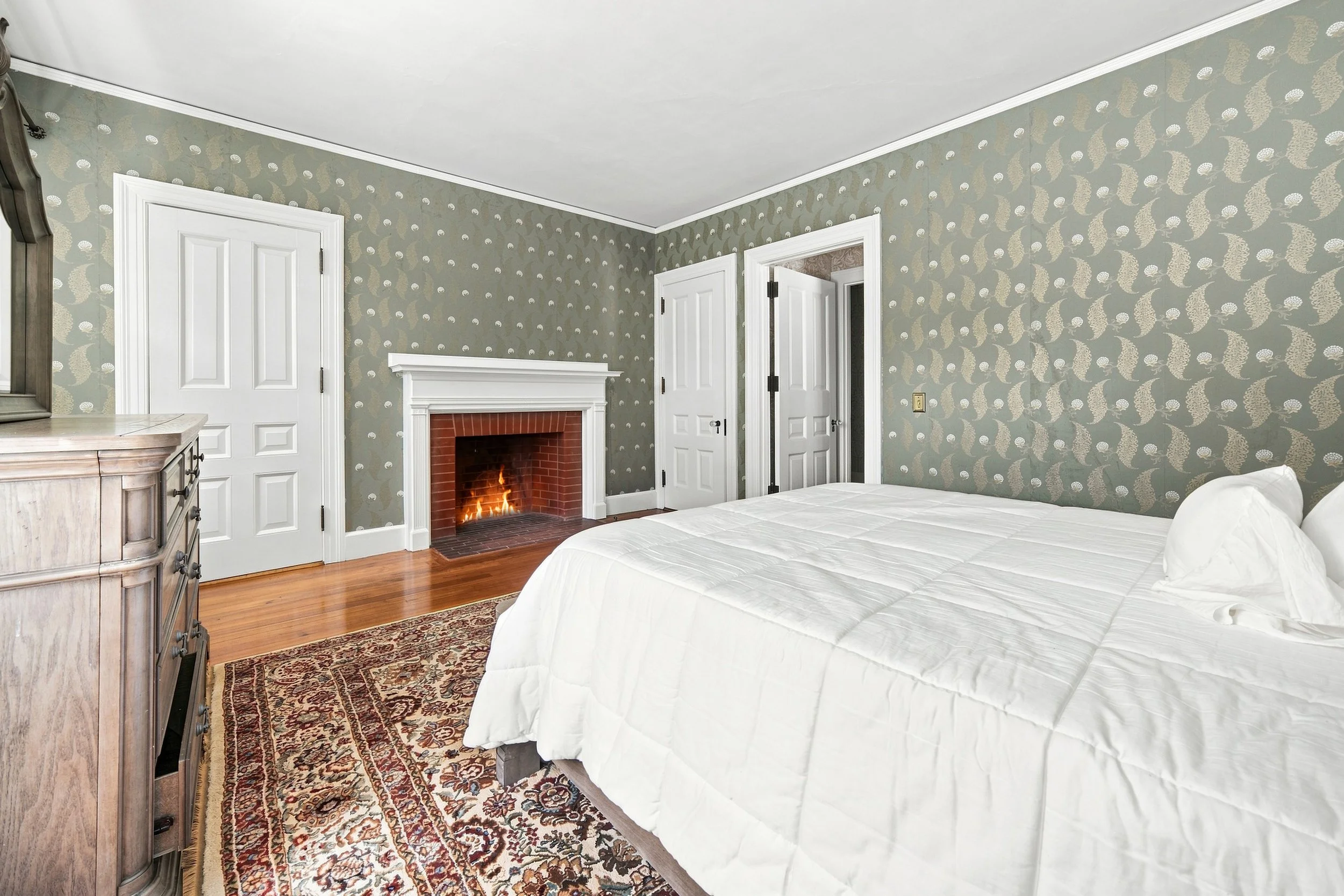 A cozy bedroom with a white bed, hardwood floor, and a fireplace with a fire burning. The walls have patterned wallpaper, and there are multiple white doors and a dresser in the room.