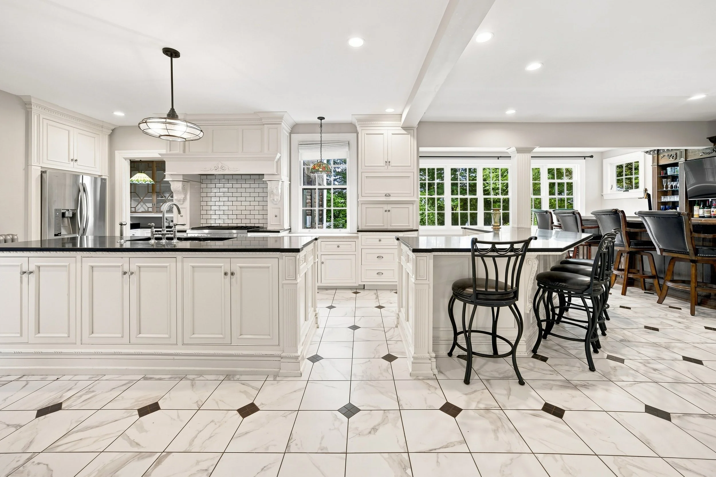 A spacious kitchen with white cabinetry, a large island with black countertop, white subway tile backsplash, and a dining area with black chairs. Natural light streams in through large windows with green trees outside.