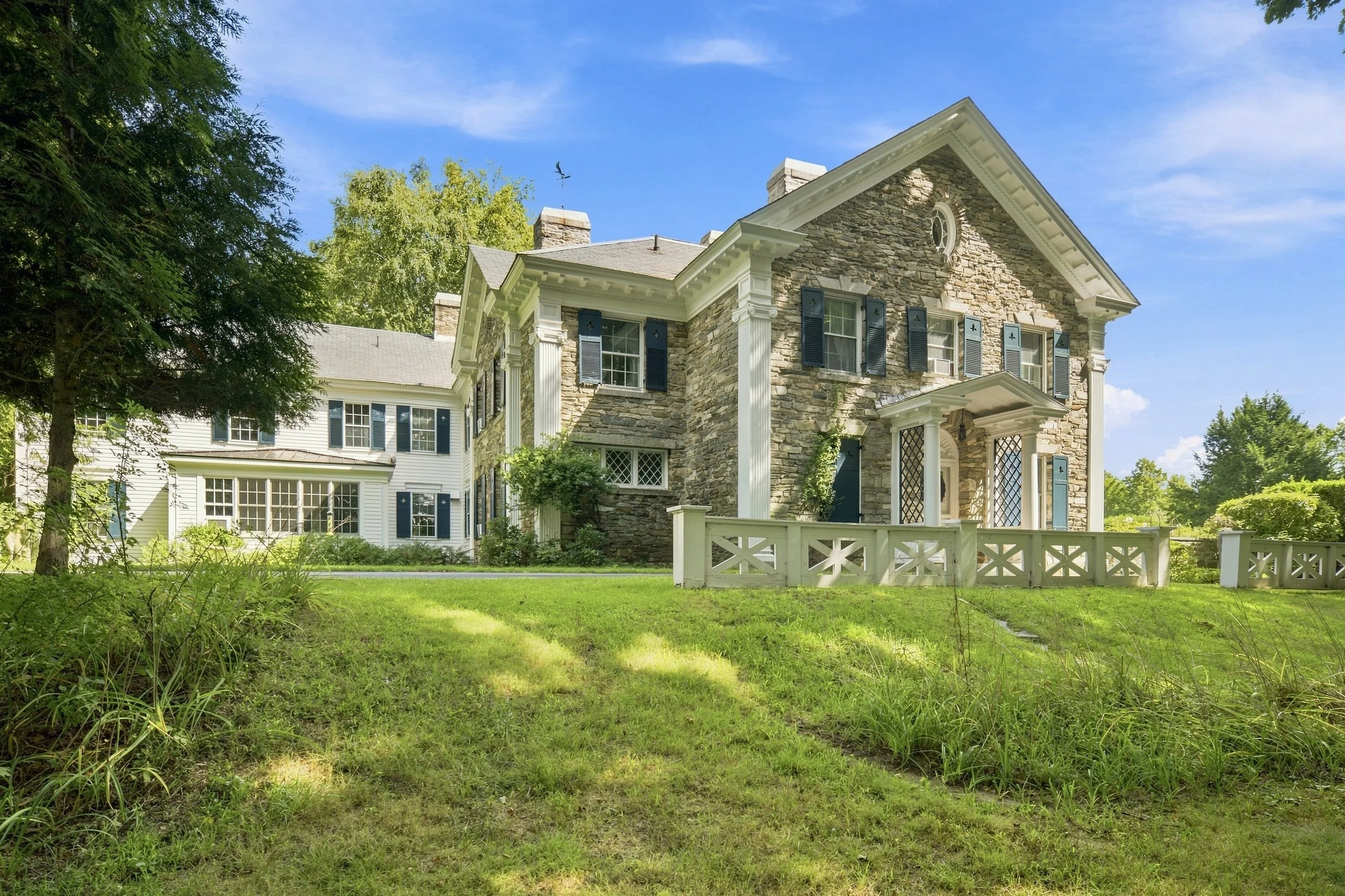A large house with stone walls and a gabled roof, surrounded by a lush green yard with trees and grass. The house has black shutters, white columns, and a small porch.