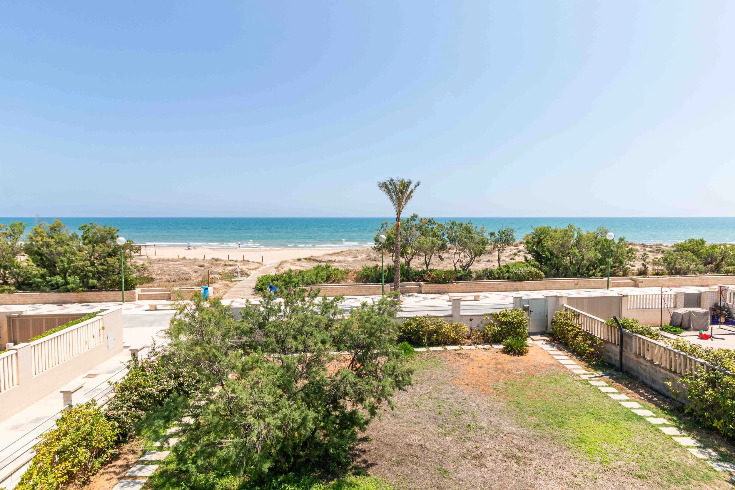 Vista de una playa con arena y el mar bajo un cielo despejado, con árboles y jardines en primer plano.