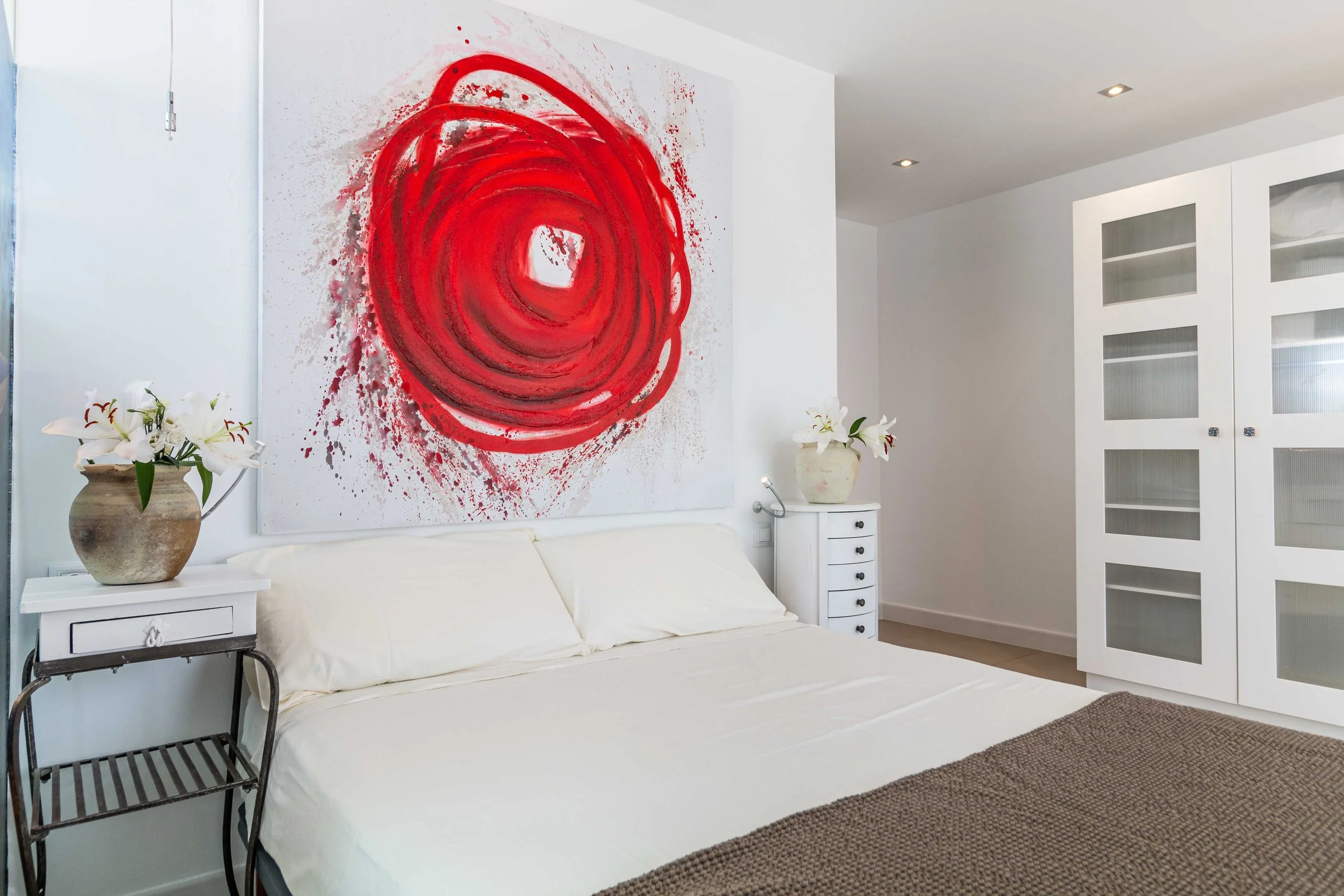 Habitación moderna con cama blanca, mesa de noche con florero y pintura abstracta roja en la pared.
