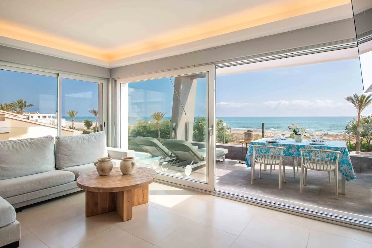 Sala de estar con ventanas grandes que ofrecen vista al mar y la playa, con muebles blancos y mesa de madera, y balcón con mesa y sillas, rodeado de palmeras y vegetación costera.