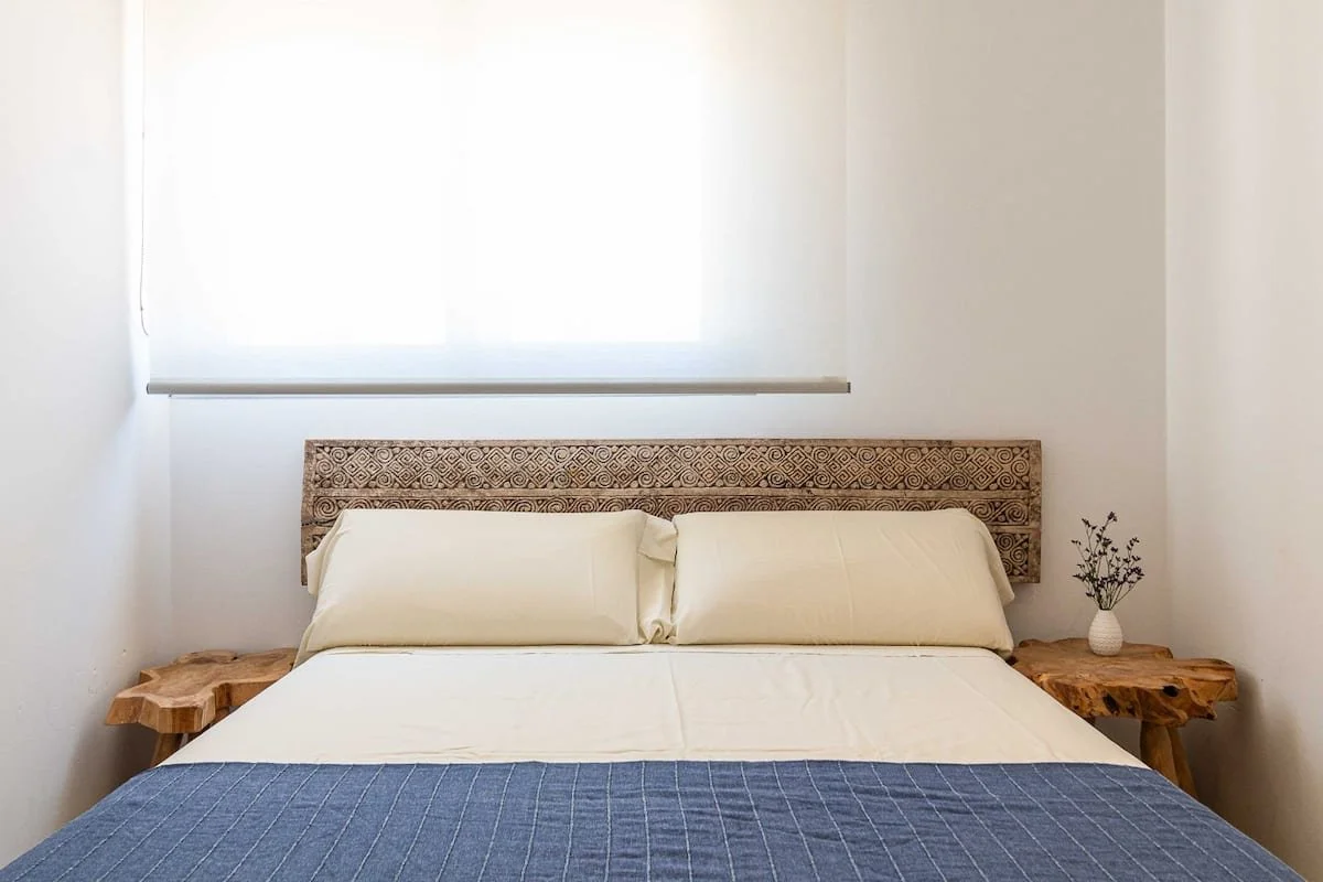 Cama con sábanas blancas y dos almohadas, cabecera de mimbre con diseño geométrico, mesitas laterales de madera rústica con planta en un jarrón, ventana con cortina blanca