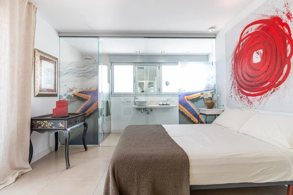 Habitación moderna con cama, arte abstracto rojo en la pared, cortinas beige, y área de baño con ducha y ventana grande.