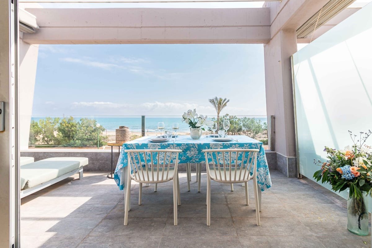 Mesa con mantel azul con flores blancas, cuatro copas de vino, platos y un florero con flores blancas, con vista al mar, en un balcón con vista panorámica, decorado con plantas y flores.