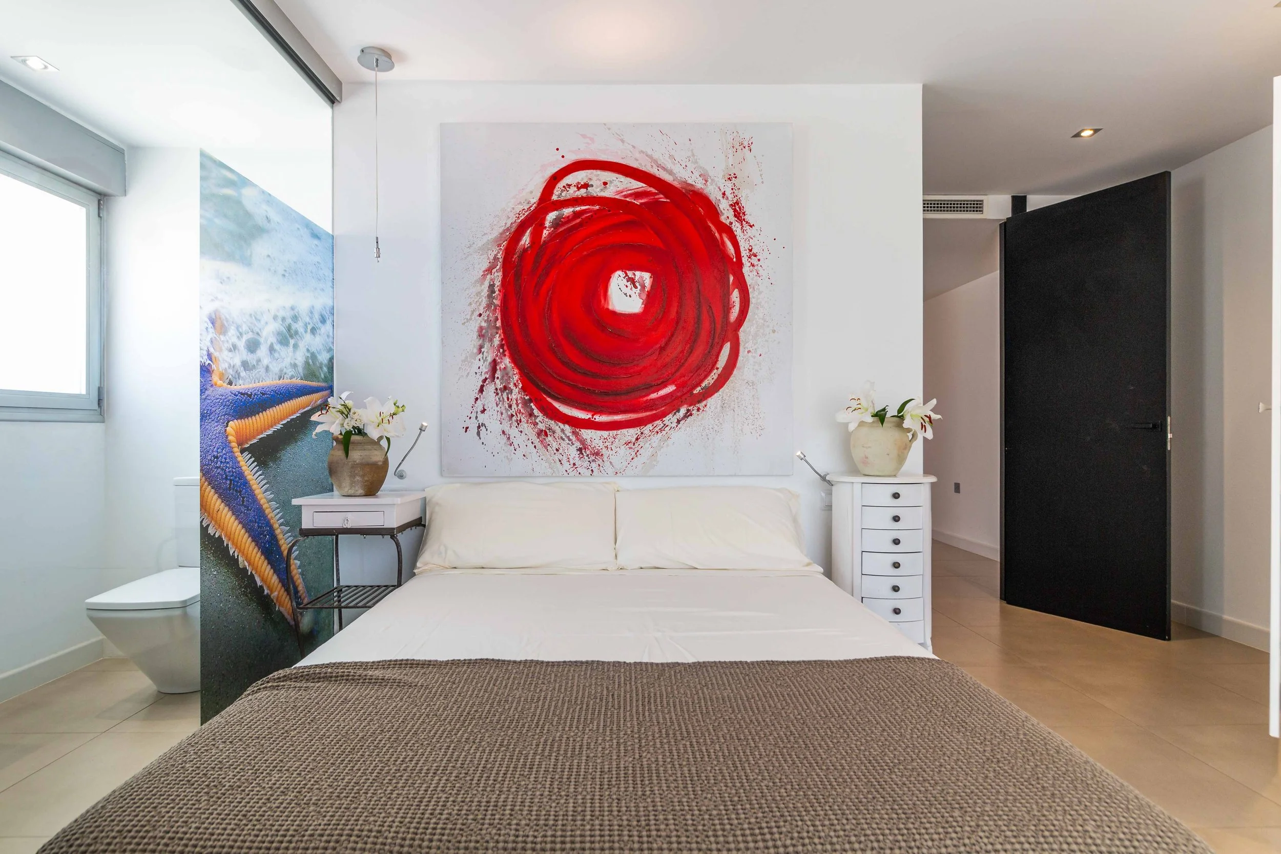 Vista de una habitación moderna con decoración en tonos claros, tocador con dos jarrones con flores blancas, y un cuadro abstracto en la pared, con una cama al frente y ventanas que permiten entrada de luz natural.