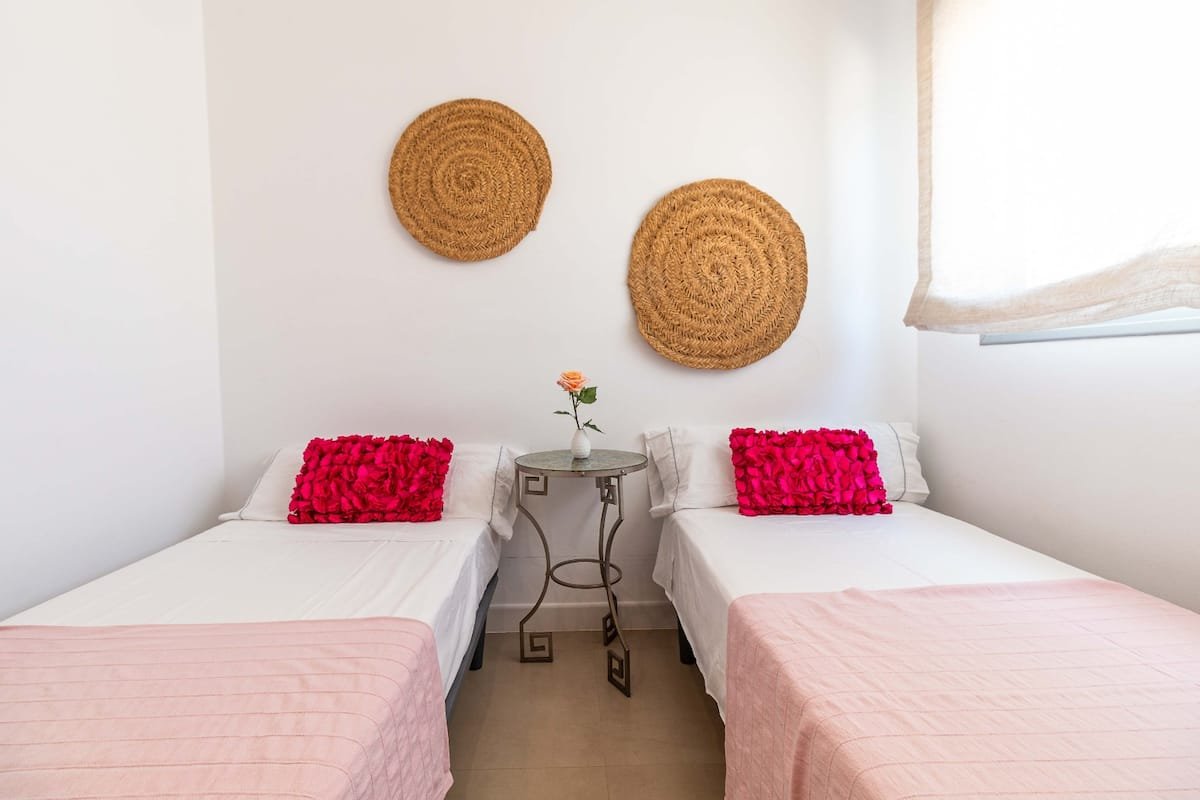 Habitación con dos camas individuales, cada una con una almohada rosa decorativa, entre ellas una mesa pequeña con una rosa en un jarrón, en la pared hay dos decorativos de palma tejidos en color marrón, y una ventana con cortina blanca