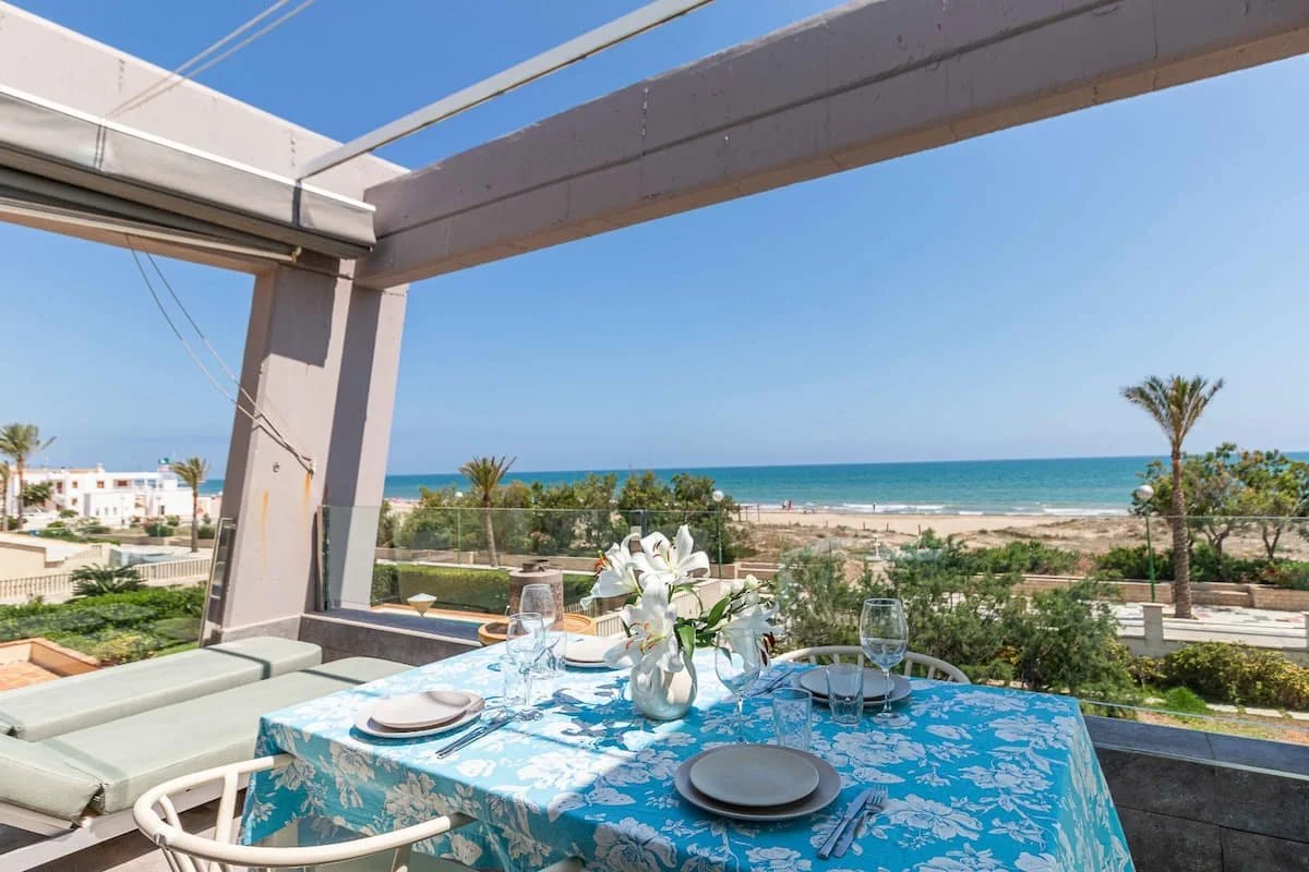 Mesa con mantel azul y flores blancas en un restaurante junto a la playa, vista del mar y palmeras en el fondo.