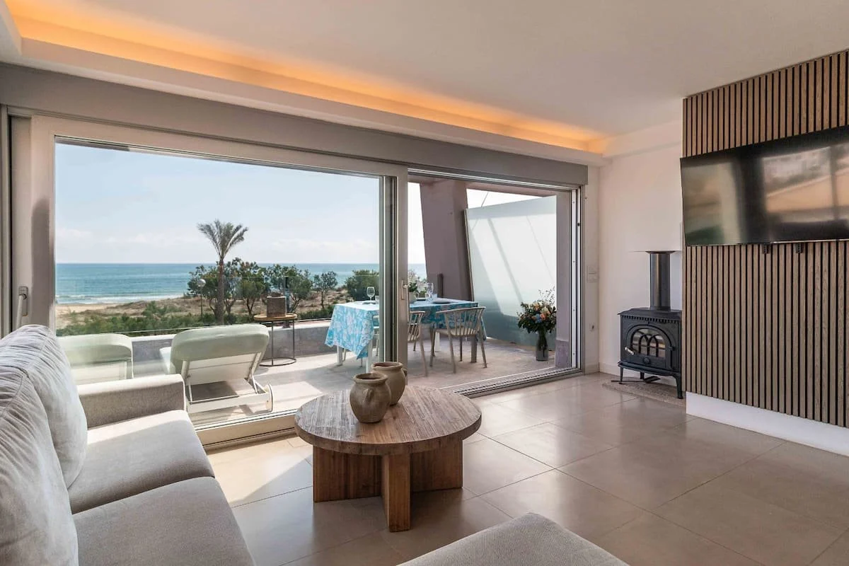 Sala de estar con vista al mar, con sofá claro, mesa de centro de madera y balcón con mesas y sillas, y vista al mar y palmeras.