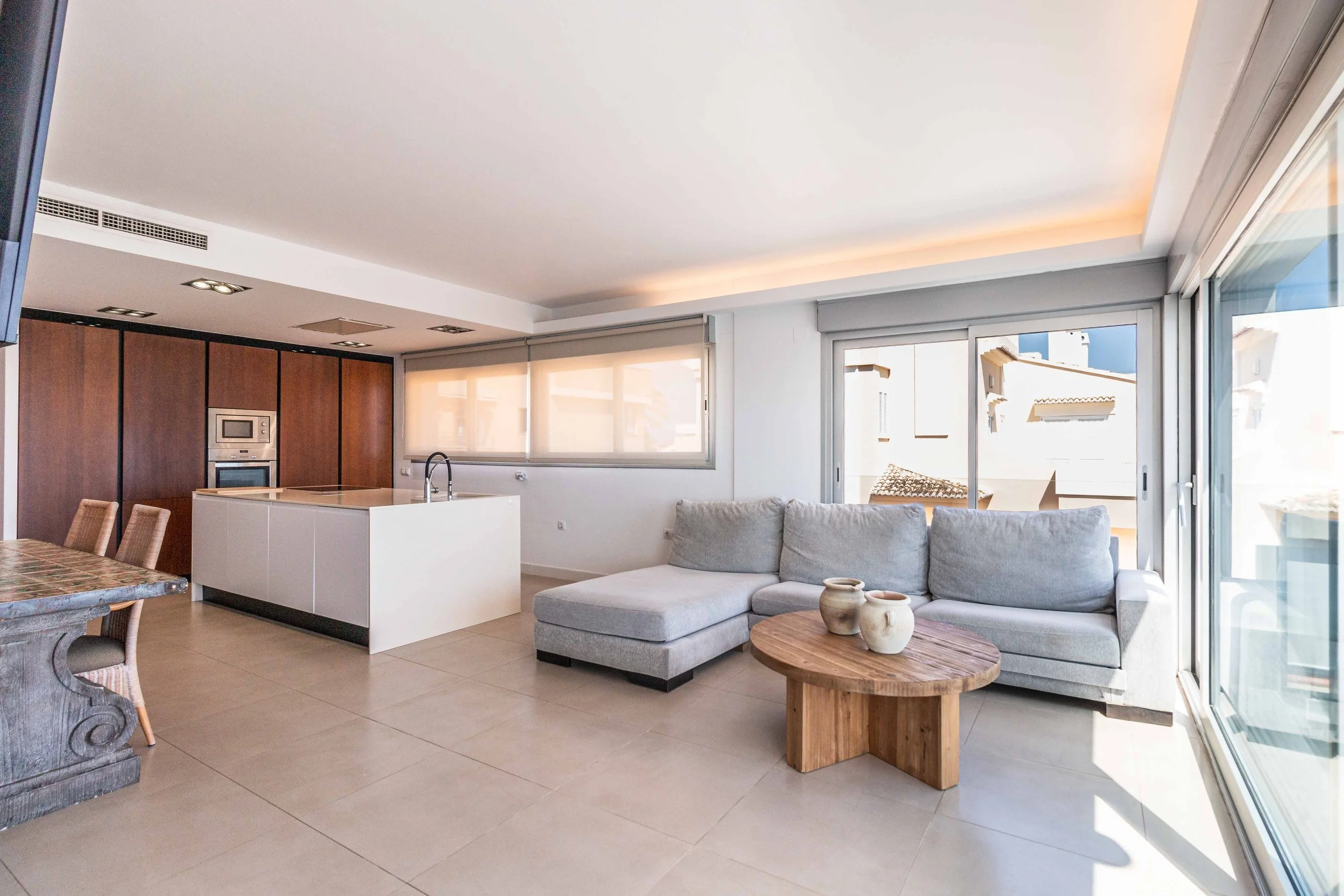 Interior de una sala de estar con sofá gris, mesa de madera, ventanas grandes y cocina moderna al fondo.