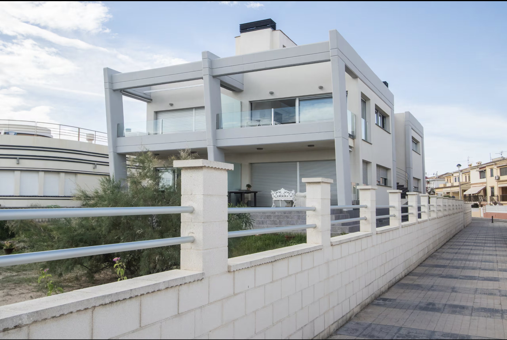 Residencia moderna de dos pisos con balcones de vidrio y una cerca blanca con barandilla metálica en un barrio residencial.