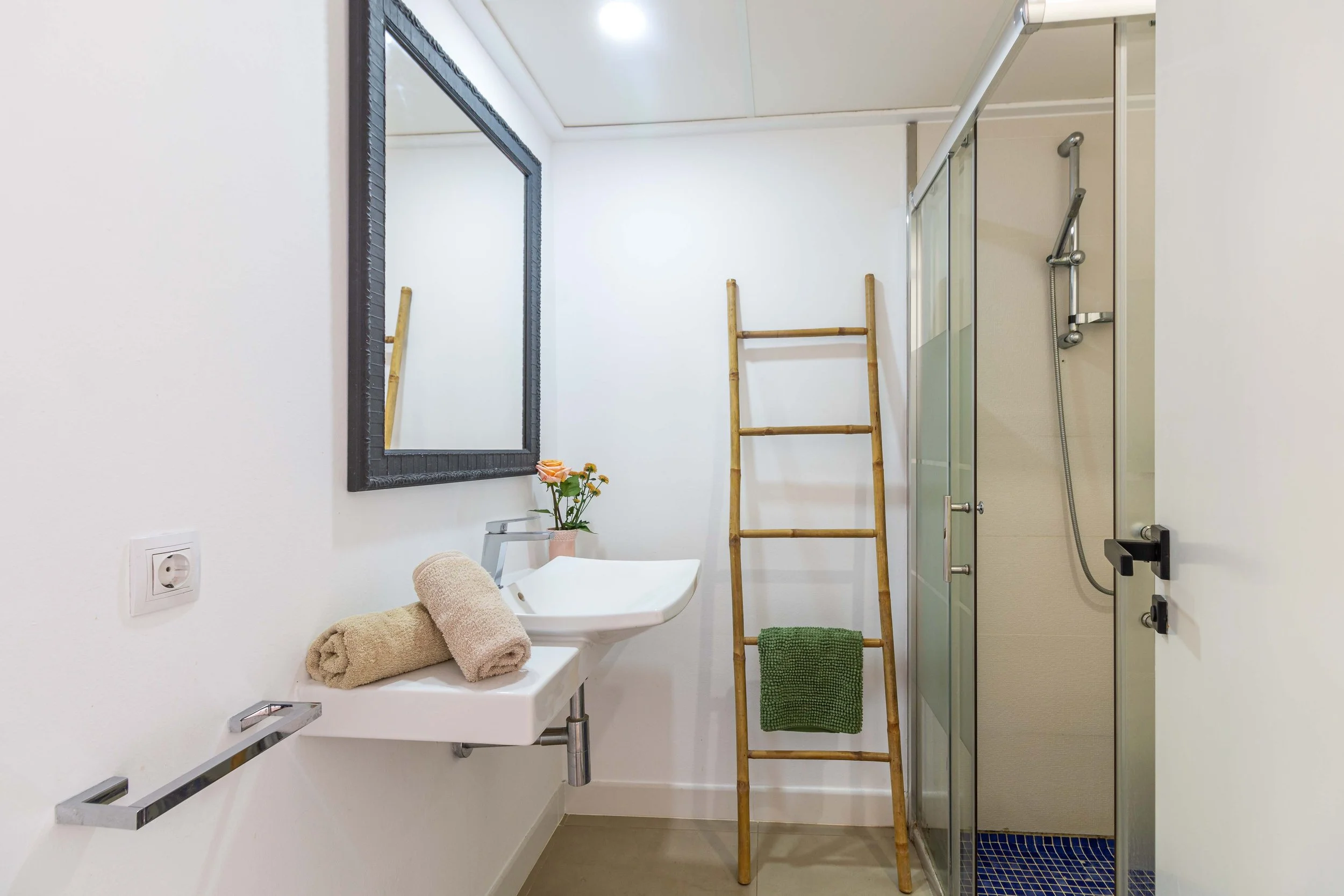 Baño moderno con espejo grande, lavabo con toallas apiladas, ducha con cristal, escalera de bambú, plantas y toallas verdes