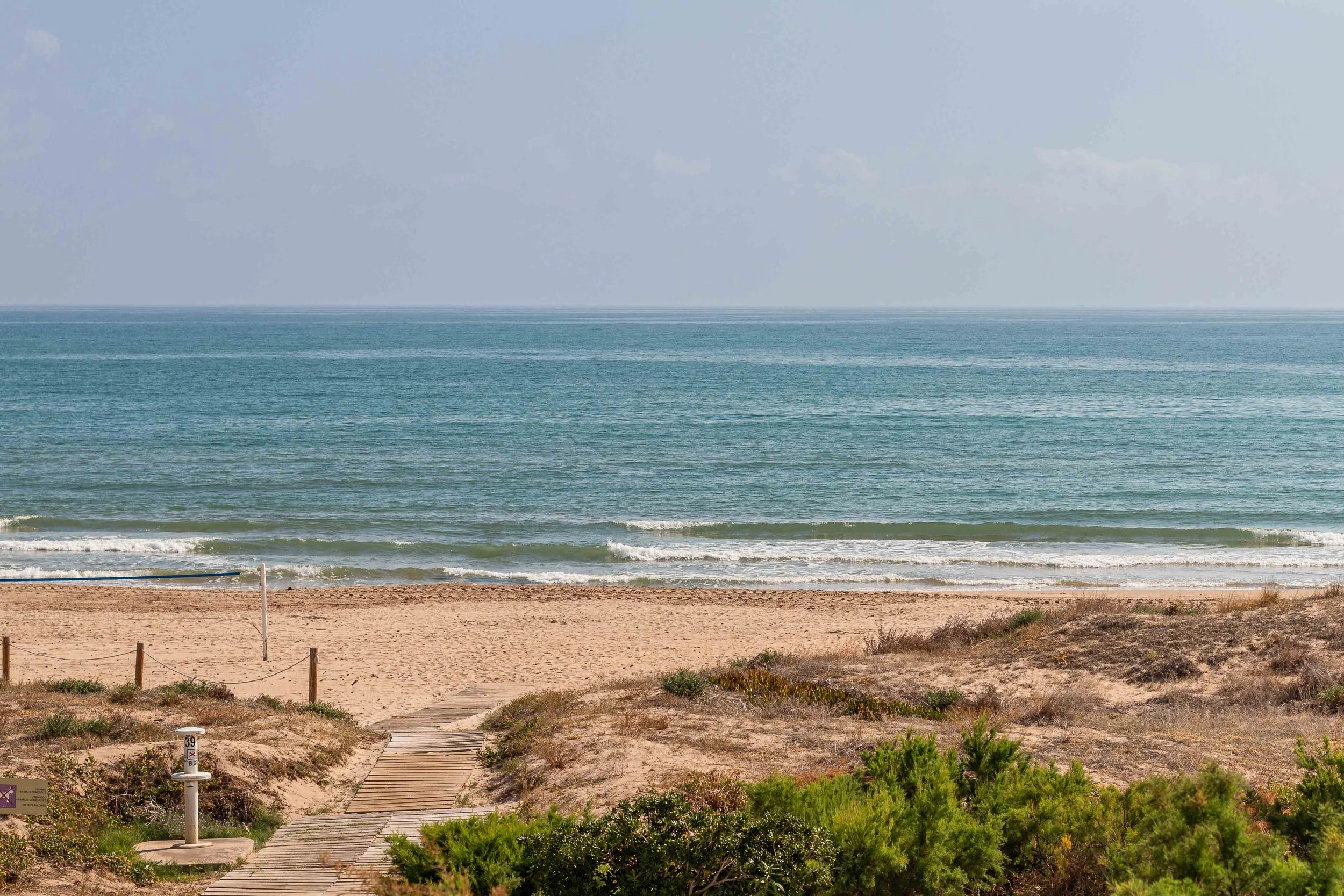 Camino de madera que conduce a la playa con arena y vegetación a los lados, vista del mar y cielo con nubes.