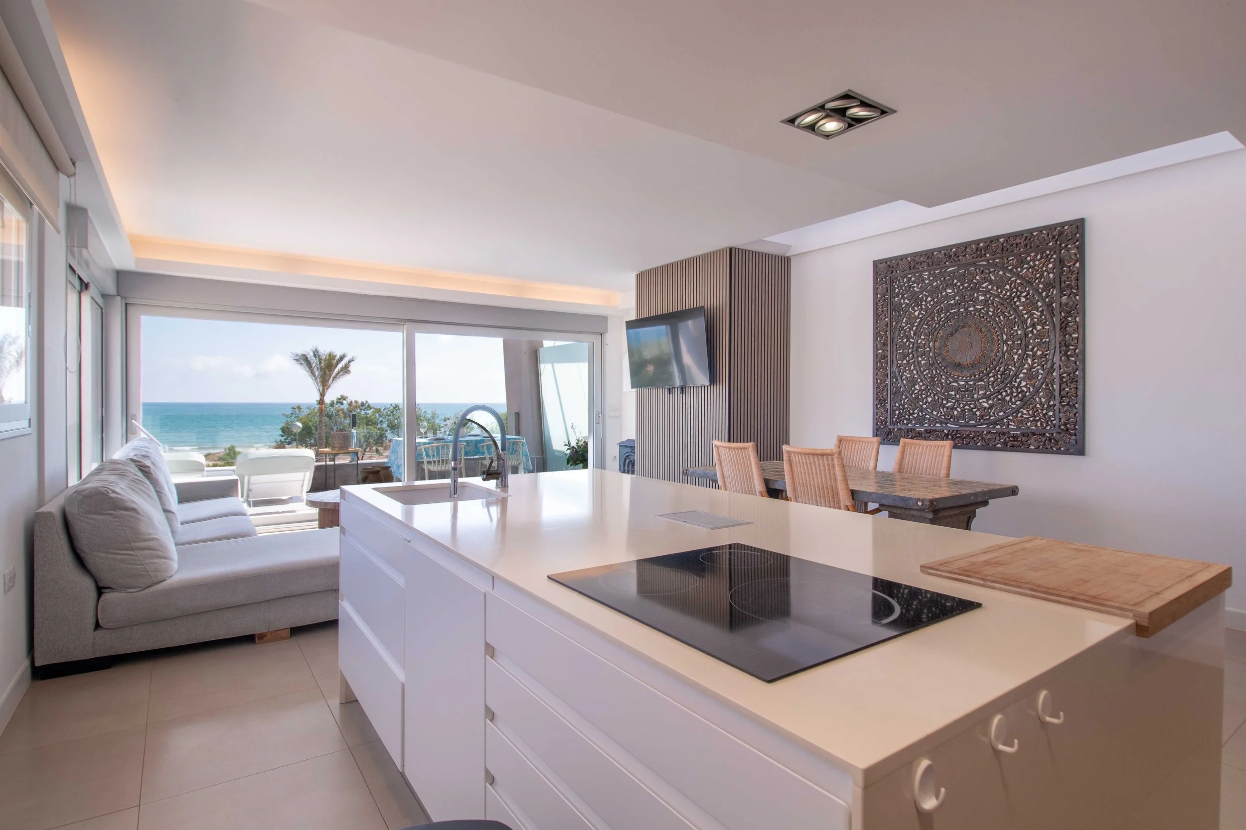 Interior de una sala con vista al mar, sofá gris, mesa de cocina blanca con cocina eléctrica, pared con decoración de madera y pantalla plana, vista al exterior con palmera y mar