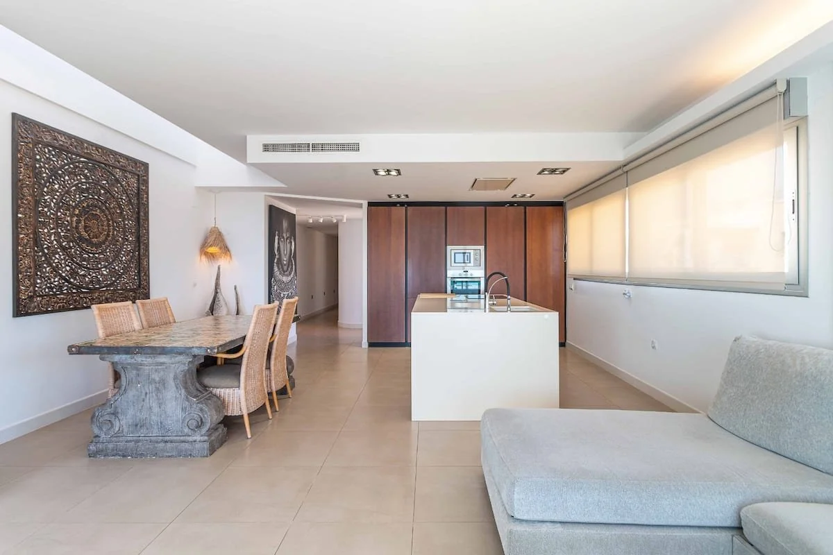 Interiores de una sala y comedor de una casa moderna, con cocina abierta, paredes blancas, ventanas grandes con persianas, y decoración con arte en las paredes.