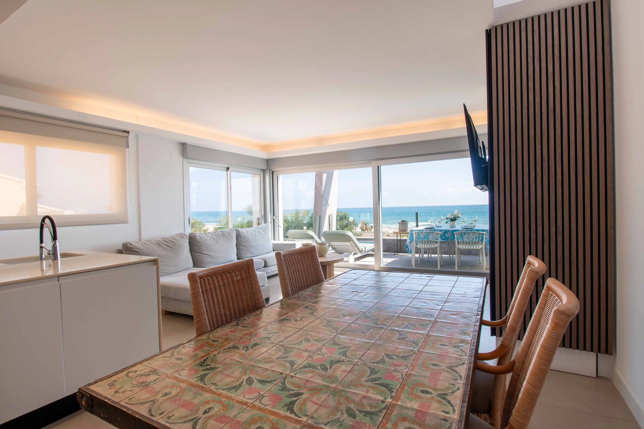 Interior de una sala con vista al mar, incluyendo una mesa con una manta de colores, silla de mimbre, sofá gris, ventanas grandes con vista al mar y un área exterior con mesa y sillas.