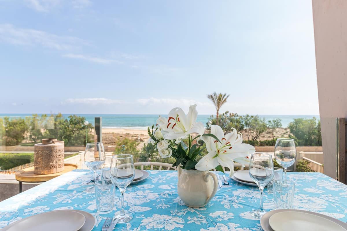Mesa decorada con un jarrón con flores lilies blancas, vasos de agua, platos y cubiertos, con vista a la playa y el mar en un día soleado