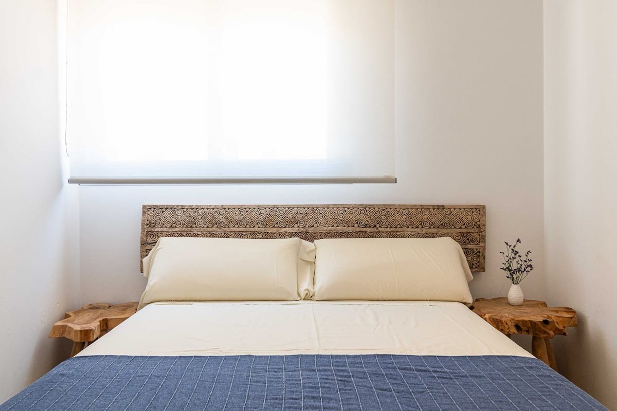 Cama con dos almohadas, cabecera de madera decorada, mesitas de noche de madera rústica y una planta pequeña en una de ellas, ventana con cortina blanca de fondo, en una habitación sencilla y luminosa.