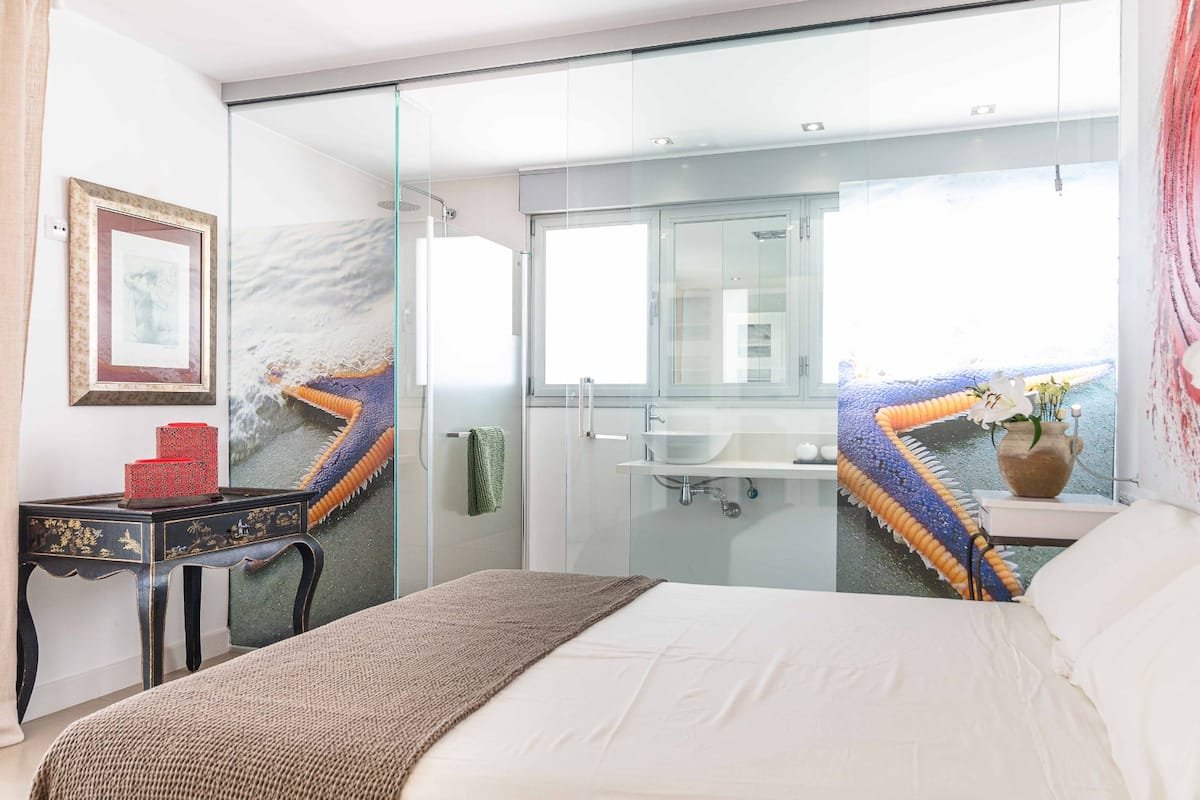Habitación con cama, en el fondo una pared con pintura que simula olas del mar y una ventana con vista a un edificio. La habitación tiene un escritorio pequeño y un lavabo, con paredes de cristal que separan el baño de la habitación.