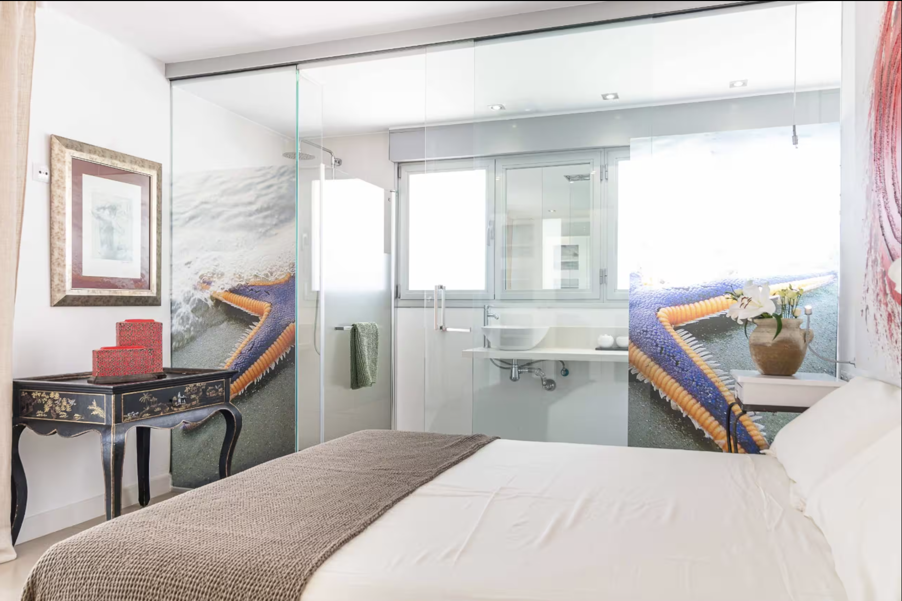 Habitación con cama, escritorio, y baño con ducha y ventana grande, decorada con arte y plantas.