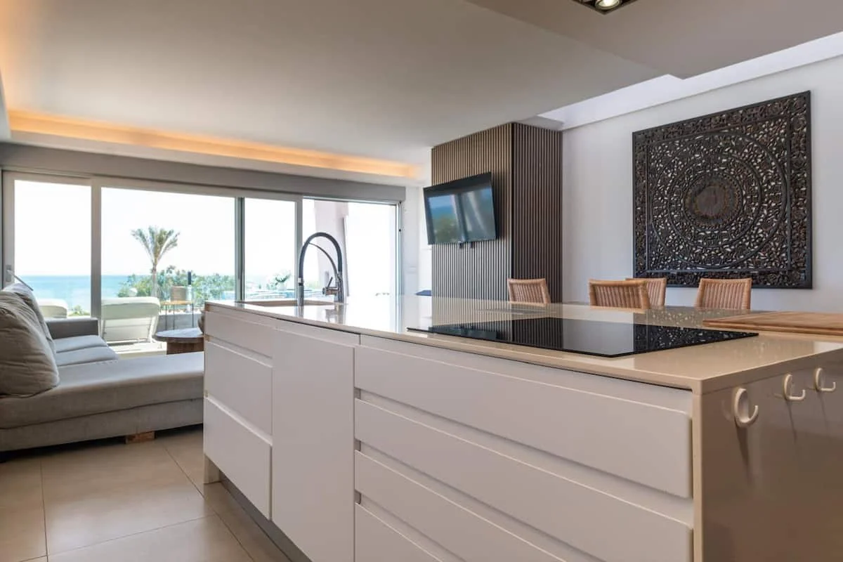 Vista de una sala y cocina moderna con vista al mar, sofá gris claro, chimenea, comedor con sillas de madera, televisión y arte en la pared.