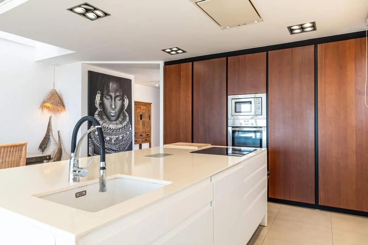 Cocina moderna con encimera blanca, fregadero y grifo negro, armario de madera con electrodomésticos integrados, y decoración artísticamente minimalista en el fondo.
