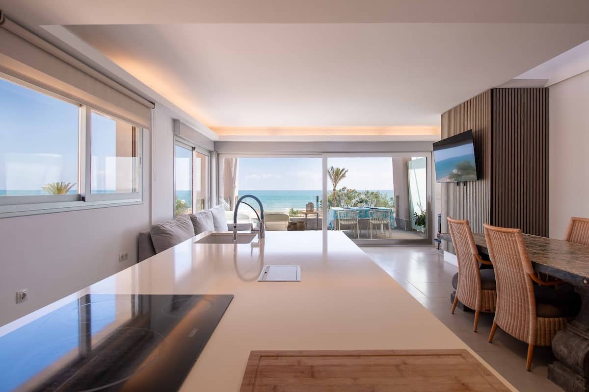 Interior de apartamento con vista al mar, sala, mesa de comedor y cocina en primer plano, terraza con sillas y mesa visible a través de ventanas y puerta corrediza