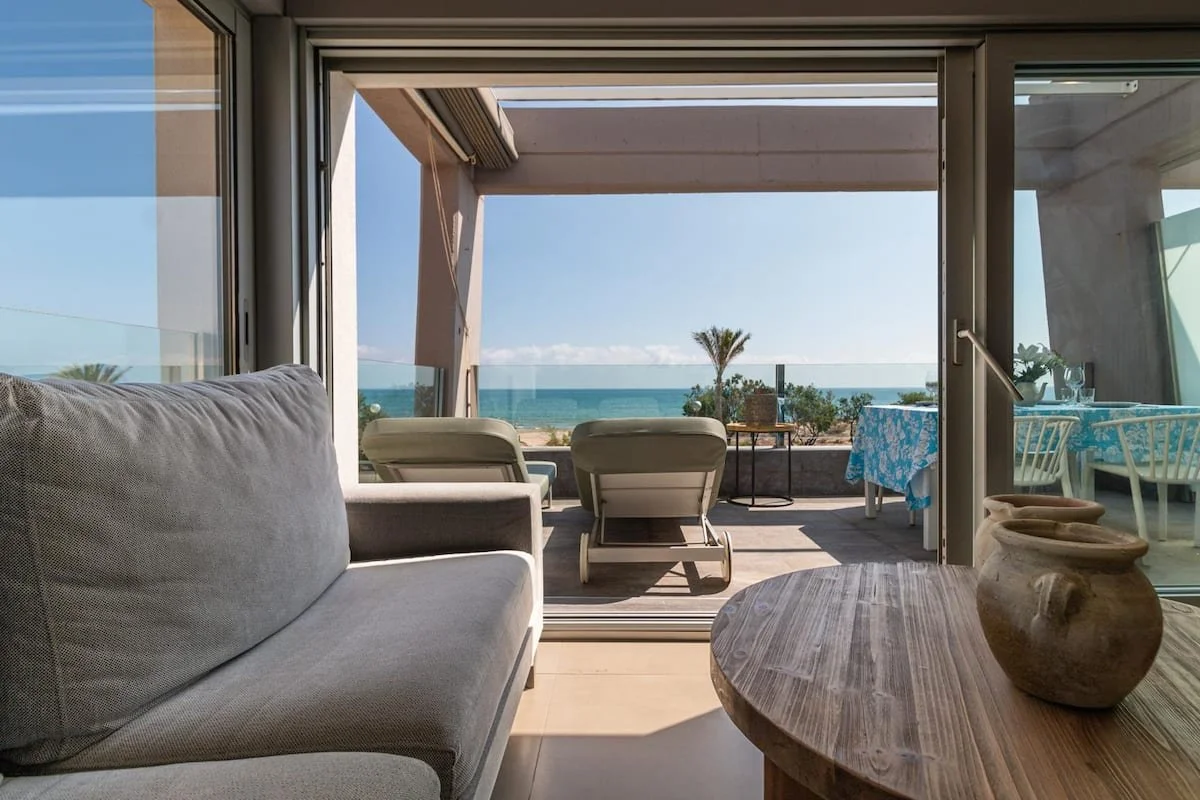 Vista de una sala con sillones y mesas desde una terraza con vista al mar y palmeras.