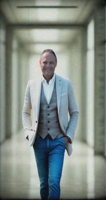 A man in a light gray blazer, white shirt, gray vest, and jeans walking in a modern corridor, smiling and with his hands in his pockets.