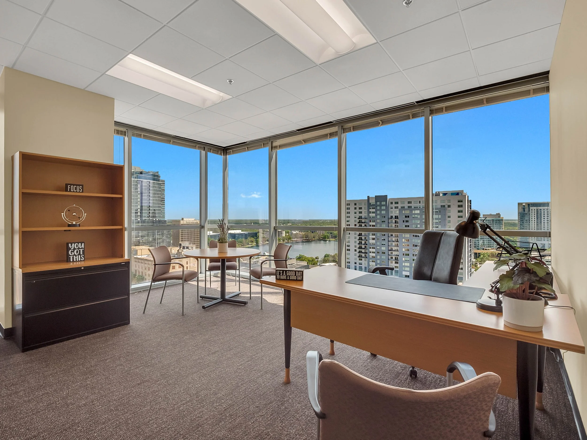 Office space with large windows showing city buildings and a river, a wooden desk with a chair, a black office chair, a bookshelf with motivational signs, a potted plant, and a bike near the window.