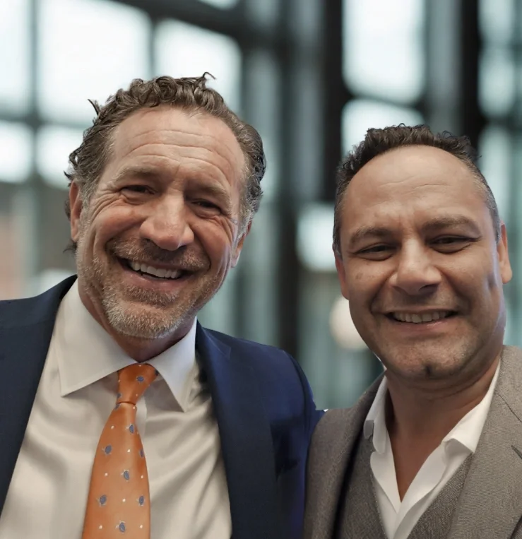 Two men smiling, dressed in business suits, indoors with a blurred modern glass building background.