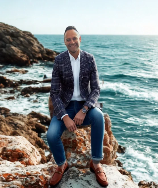 A man sitting on rocks near the ocean, smiling at the camera, wearing a plaid blazer, white shirt, jeans, and loafers.