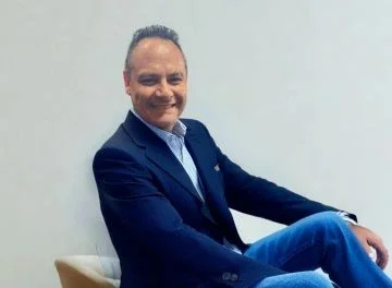 Man in a navy blazer and light blue shirt sitting on a beige chair against a plain white background.