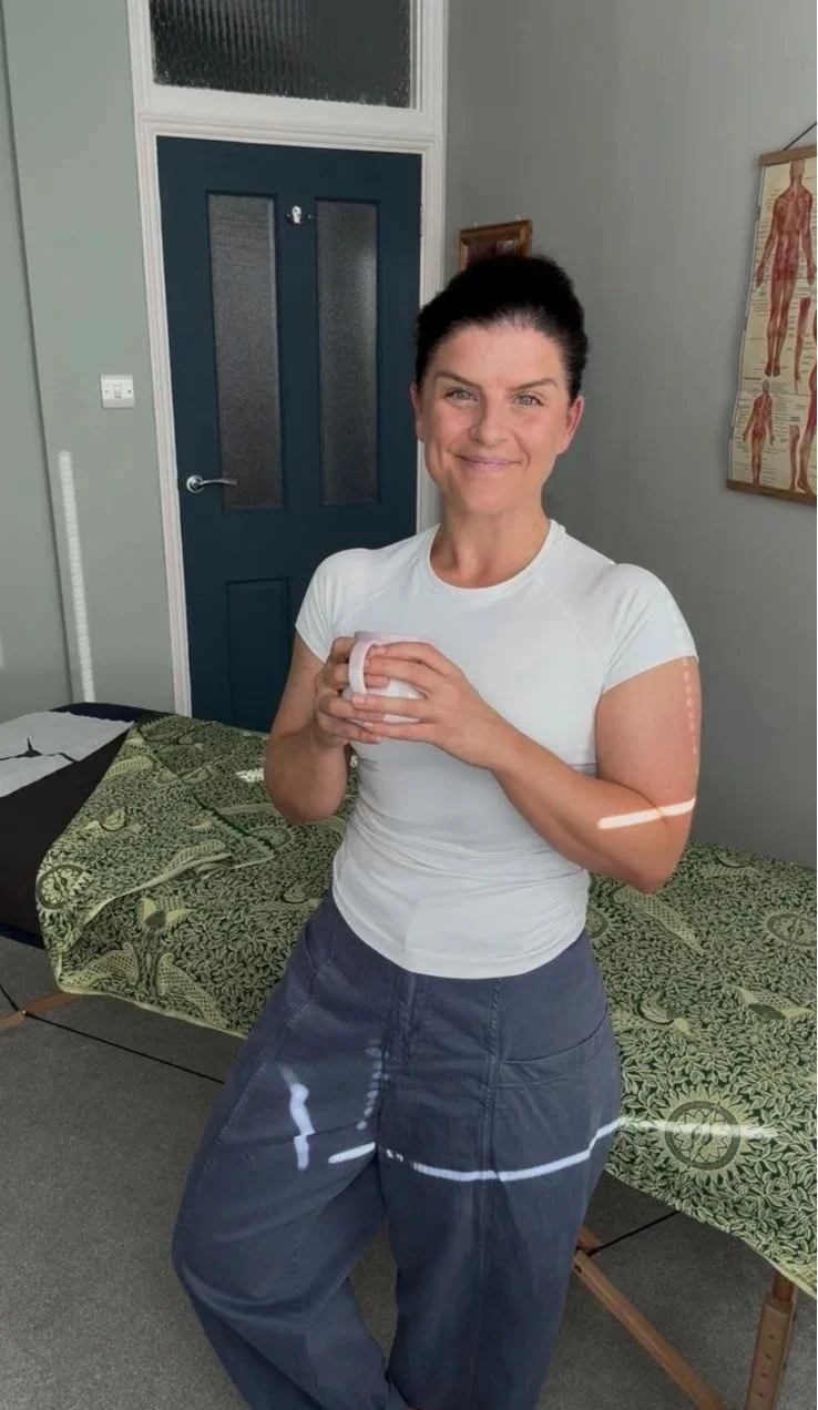 A woman smiling while holding a white mug in a room with a blue door, anatomical chart on the wall, and a bed with green patterned sheets.