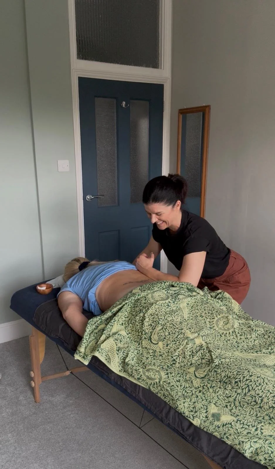 A woman receiving a massage from a massage therapist in a room with a closed blue door, a mirror, and light-colored walls.
