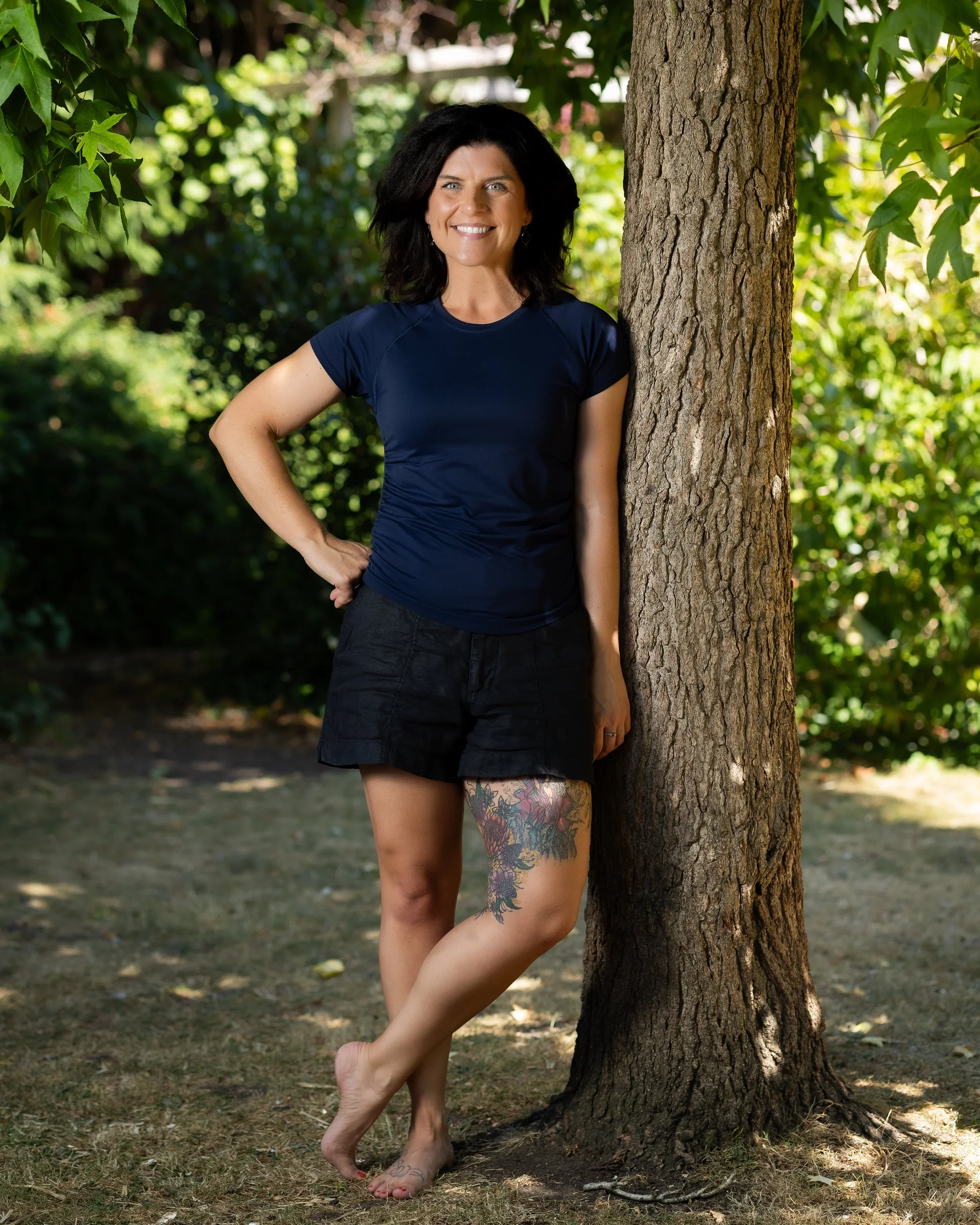 A woman with dark hair and tattoos on her thigh, wearing a navy blue shirt and black shorts, standing outdoors next to a tree with green foliage in the background.