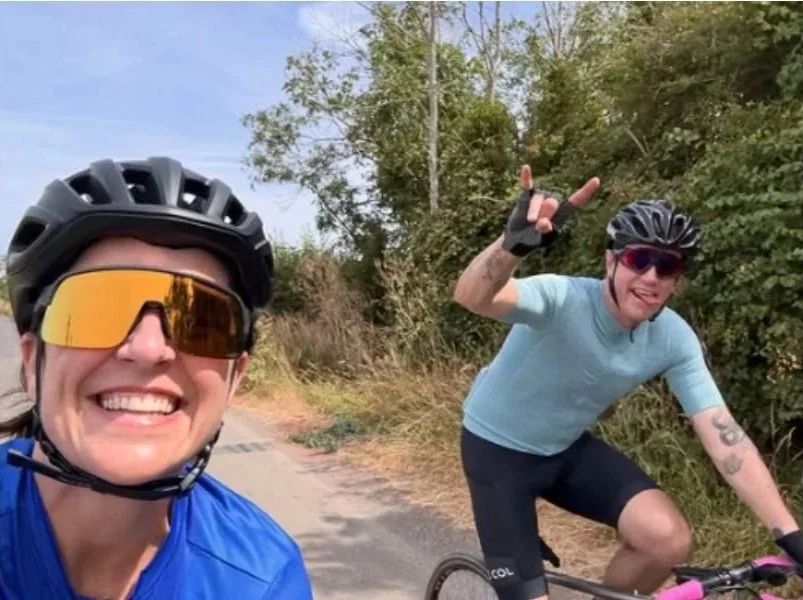 Two women cycling outdoors on a sunny day, wearing helmets and sunglasses, with one smiling at the camera and the other making a hand gesture.