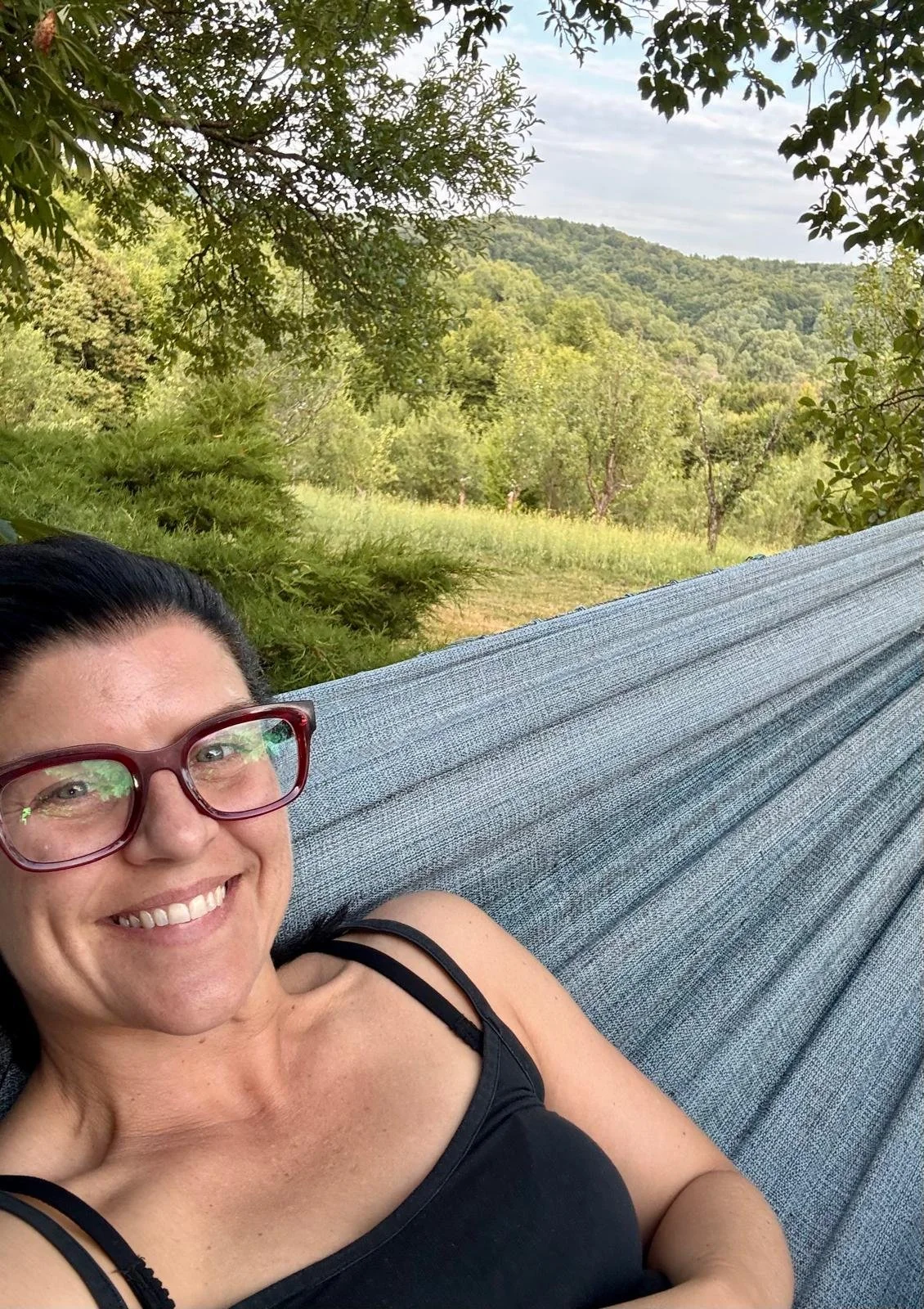 A woman with red glasses smiling and lying in a hammock outdoors, with green trees and mountains in the background.
