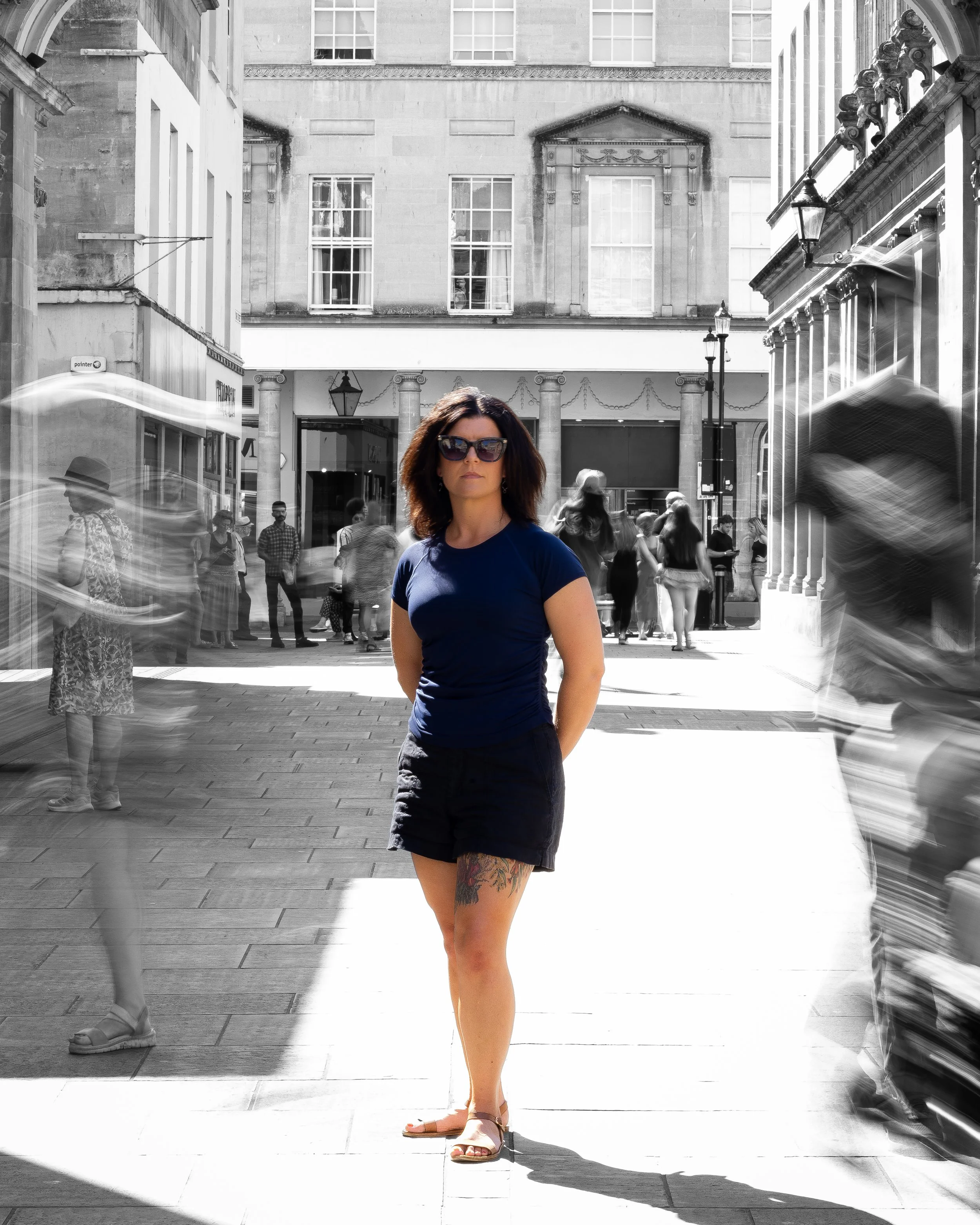 A woman with brown hair wearing sunglasses, a blue t-shirt, black shorts, and sandals standing on a city sidewalk. The background features a crowd of people, historic buildings with large windows, and street lamps. The photo has been edited to be mostly black and white, with the woman in color.
