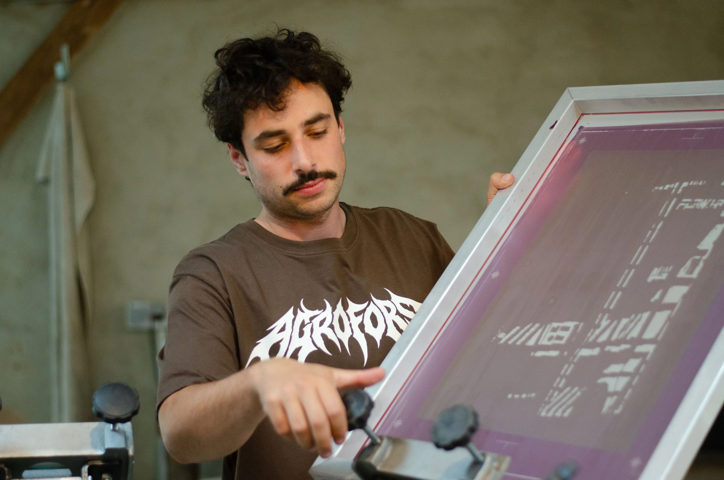Mann mit lockigem Haar hält in einem Atelier einen großen Siebdruckrahmen mit violettem Druckmotiv.