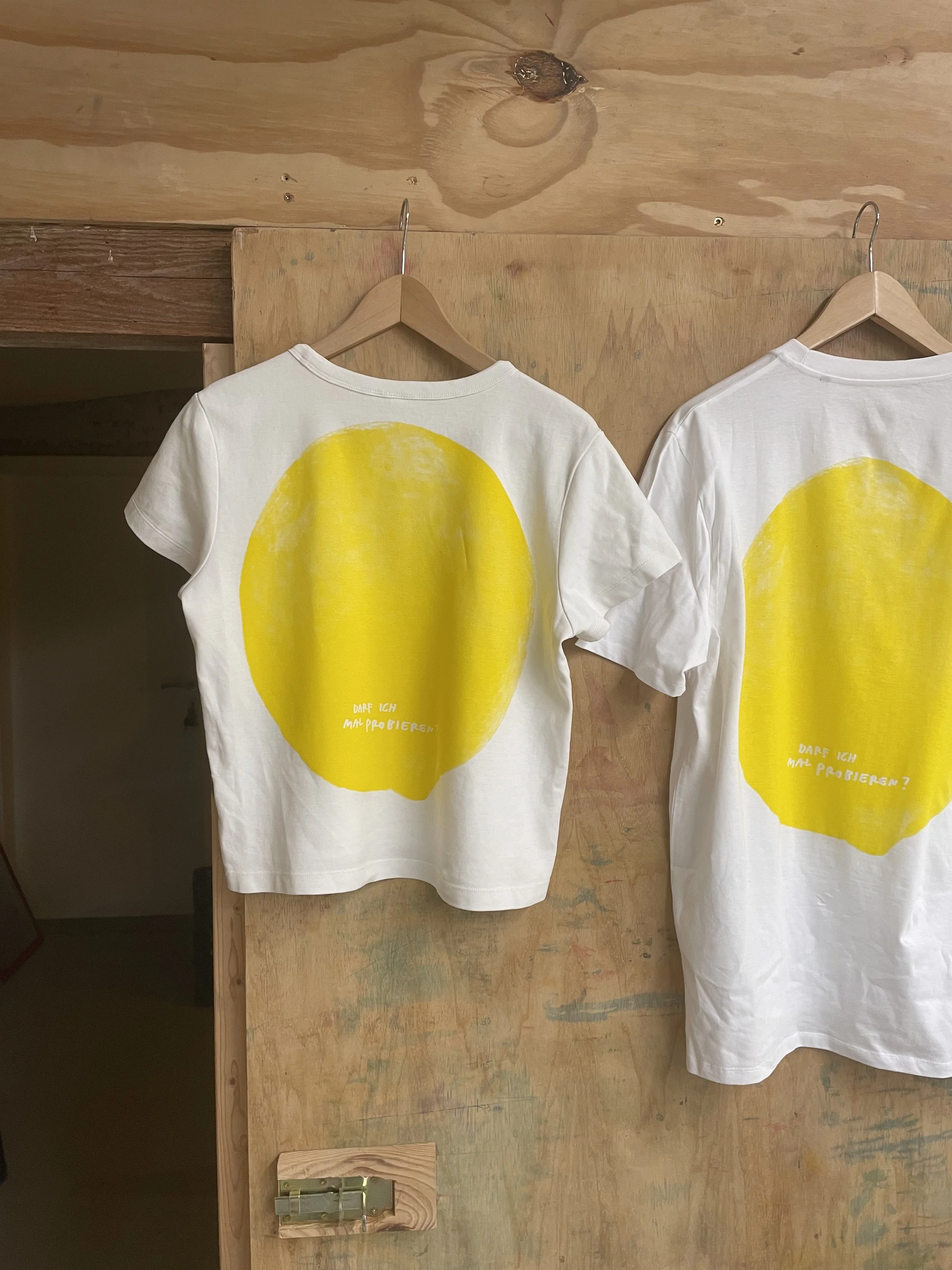 Zwei weiße T-Shirts mit großen gelben Zitronendrucken hängen auf Holzbügeln an einer rustikalen Holzwand.