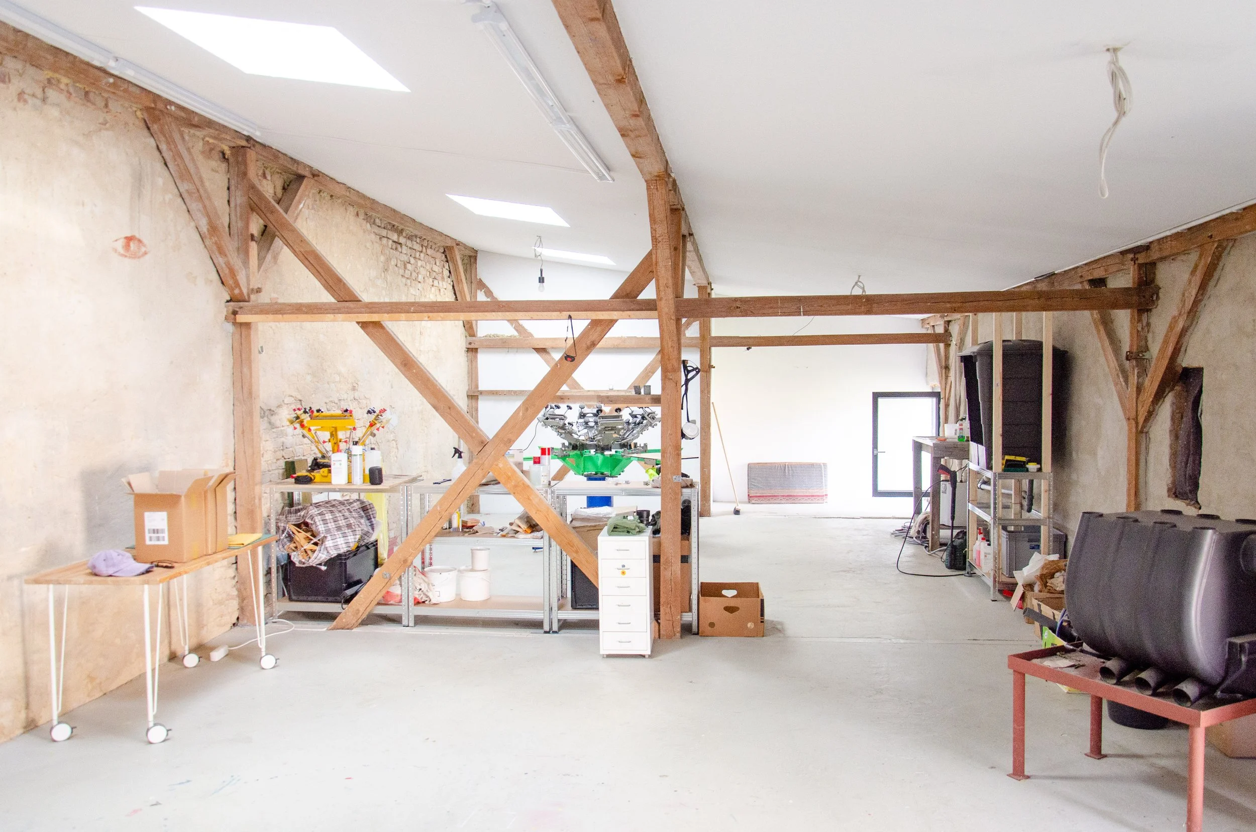 Helle Werkstatt mit sichtbaren Holzbalken, Siebdruckmaschine, Regalen, Werkzeugen und Lagerkartons