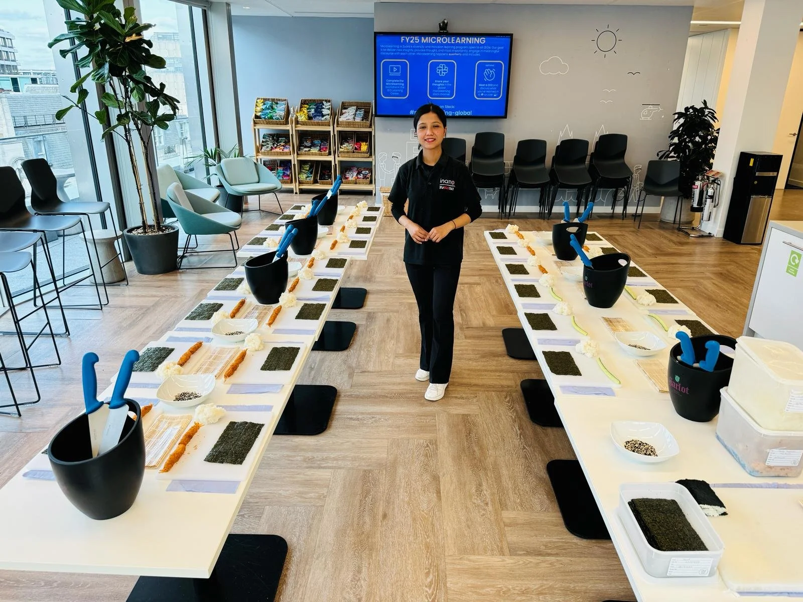 A smiling woman stands in a classroom or workshop setting with long tables set up for a sushi-making activity. The tables have prepared ingredients like seaweed sheets, rice, and utensils, with a few bowls and tools arranged neatly.