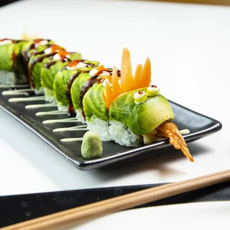 Sushi roll shaped like a Dragon on a black plate, garnished with wasabi, cucumber eyes, and carrot accents.