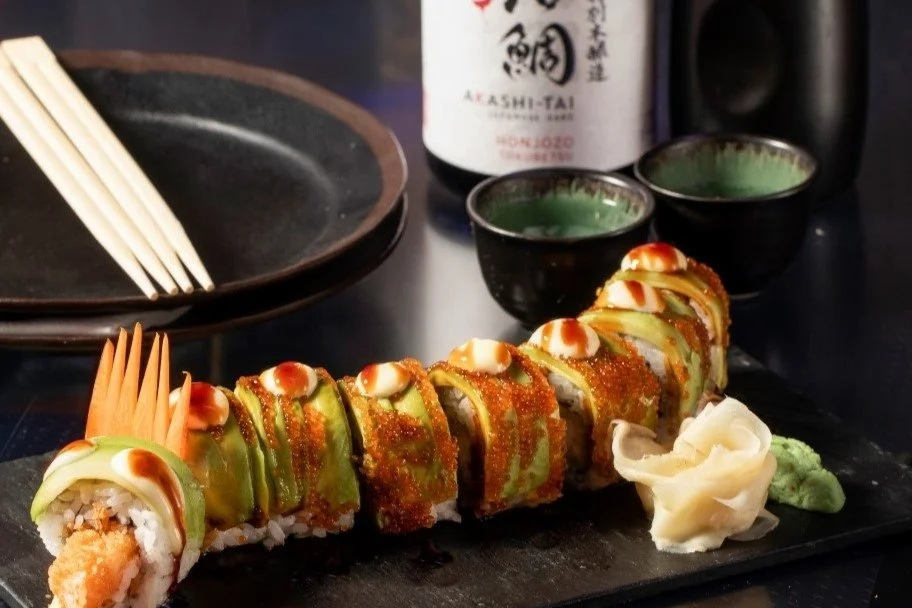 A plate of assorted sushi rolls with orange and green toppings, garnished with ginger and wasabi, with soy sauce, a black plate, chopsticks, and two small cups of sake in the background.