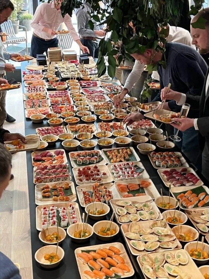 A large table filled with an assortment of sushi and Japanese dishes, with people serving themselves at a buffet-style gathering.