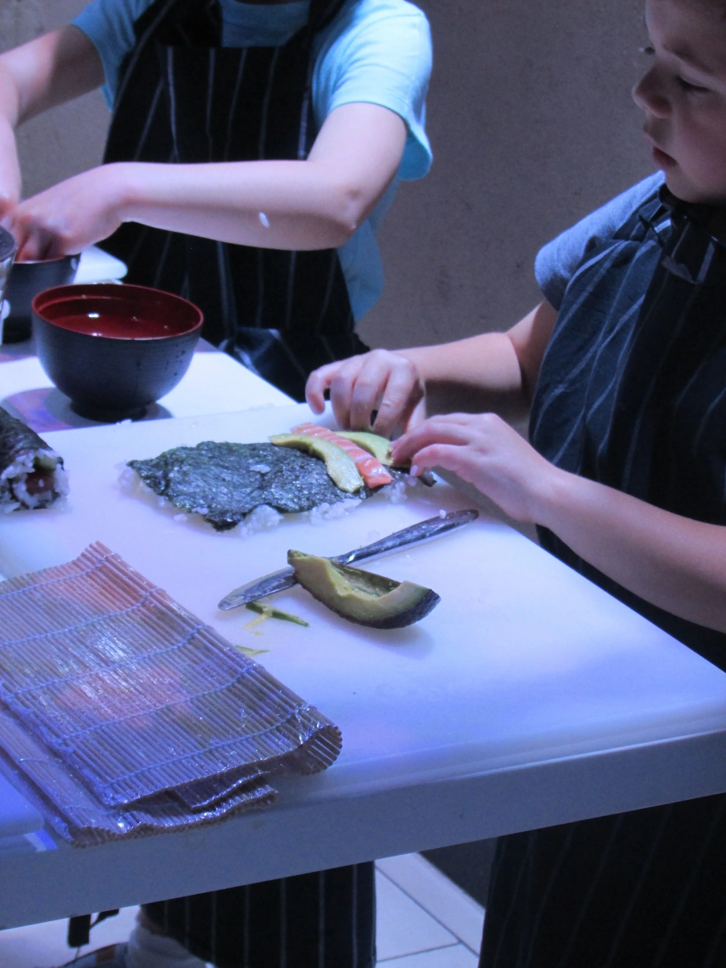 Children preparing sushi in a dimly lit room, with seaweed, sushi rolls, rice, avocado, and a knife on the table.