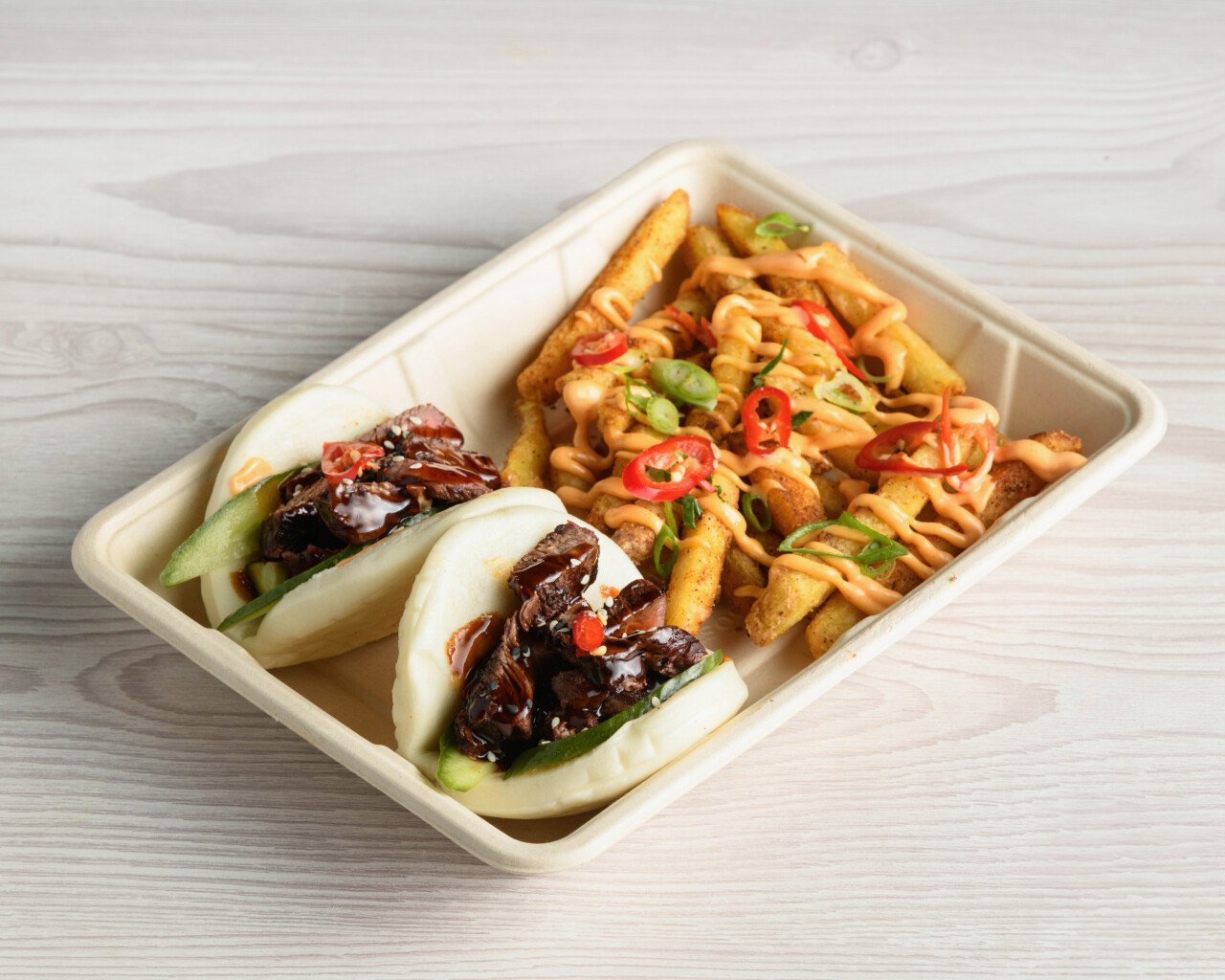 Beef Teriyaki bao buns with fresh cucumber and sauce, alongside loaded fries drizzled with spicy mayo and topped with sliced red chili and green onions.