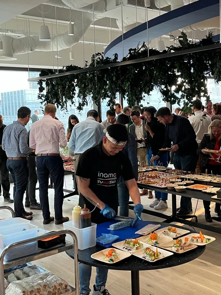A chef live rolling sushi at a table during a buffet in a modern, well-lit room with large windows, surrounded by many guests.